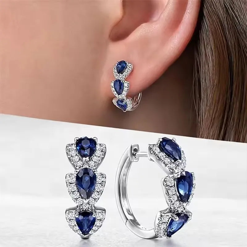 Silver hoop earrings with blue gemstones worn by a person, displayed on a white surface.